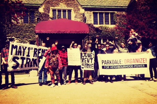 Des membres d'un mouvement de locataires se rassemblent pour une grève des loyers, tenant des pancartes avec les inscriptions "MAY 1st RENT STRIKE", "JUNE 1 RENT STRIKE", et "PARKDALE: ORGANIZE! FOR NEIGHBOURHOOD POWER!", devant un bâtiment avec une toiture en pente.
