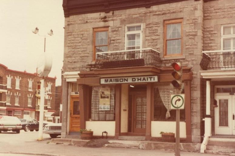 Bâtiment en pierre avec le enseigne 'MAISON D'HAITI', feux de circulation et voitures garées sur la rue.