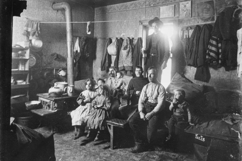 Une famille assise dans une pièce sommairement meublée, avec des murs tapissés et des vêtements suspendus. Des enfants, un homme et une femme sont présents, exprimant des rôles familiaux dans un cadre de vie modeste.