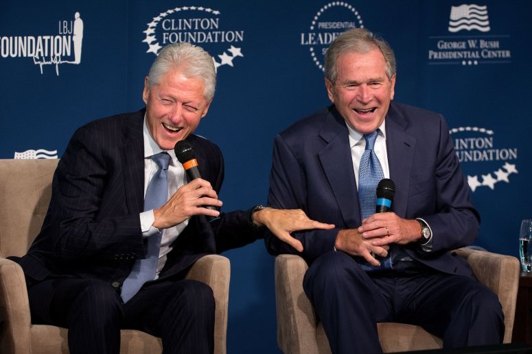 Deux hommes souriants, Bill Clinton et George W. Bush, assis sur des chaises lors d'un événement de la Clinton Foundation, avec un fond bleu affichant des logos de fondations.