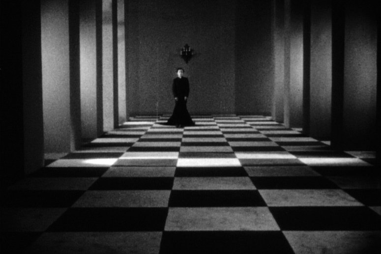 Une scène en noir et blanc présentant une femme vêtue de noir, debout sur un sol en damier, entourée de murs verticaux qui créent un effet dramatique et surréaliste.