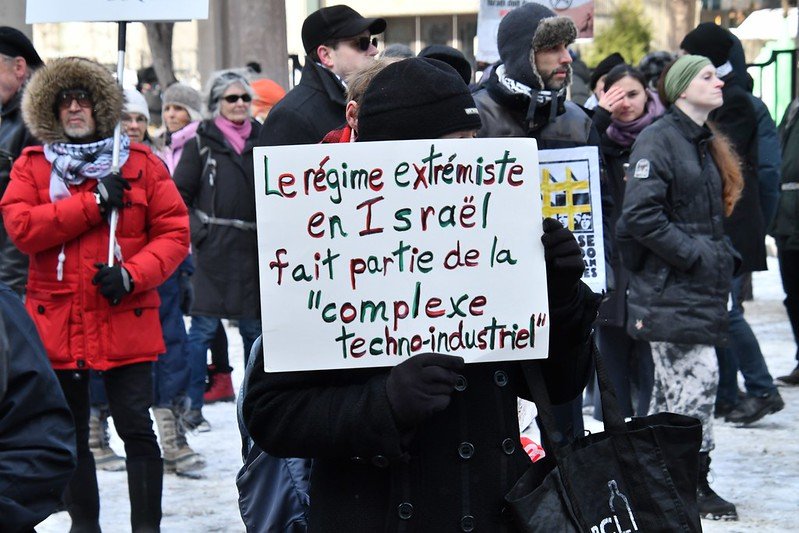 Une manifestation à Montréal pour Palestine. Des manifestants tiennent une pancarte sur laquelle il est écrit "Le régime extrémiste en Israël fait partie de la complexe techno-industriel".